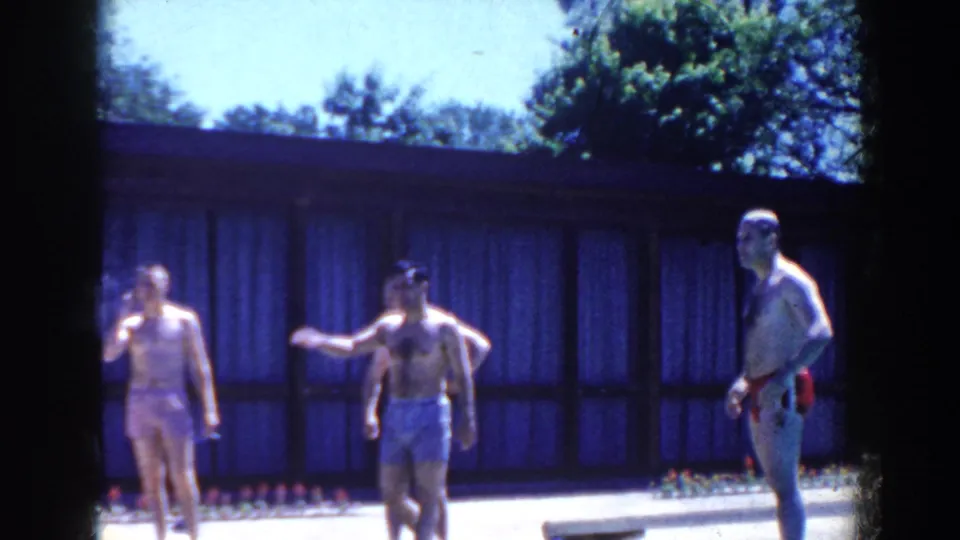 1958 French Lick frame showing adults on a pool deck, including a person with a cigarette, in front of a low building and trees.