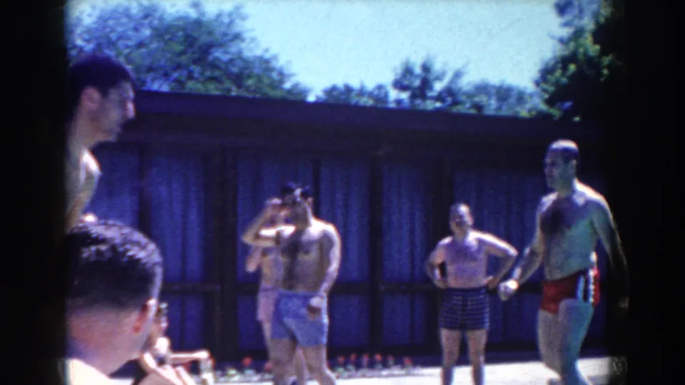 1958 French Lick home-movie frame of adults in swimsuits standing near a pool, with a low building and trees in the background.