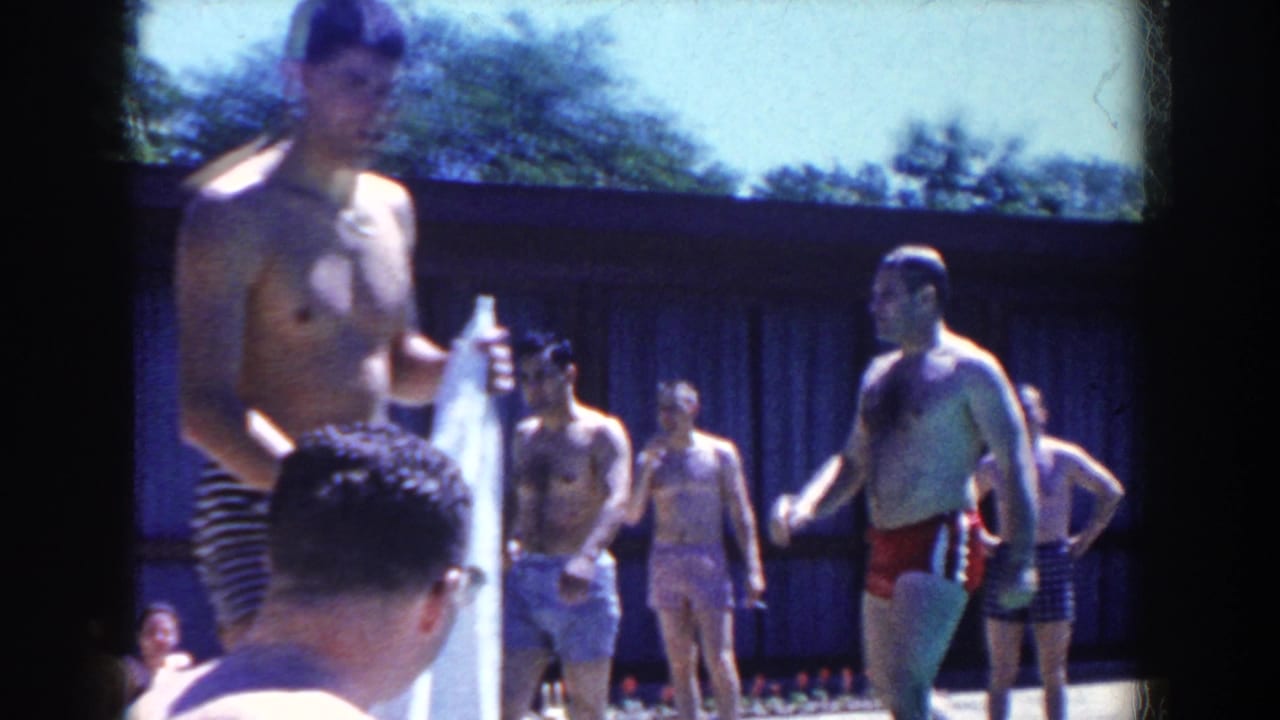 Men in swim trunks stand outdoors near a pool on a sunny day.