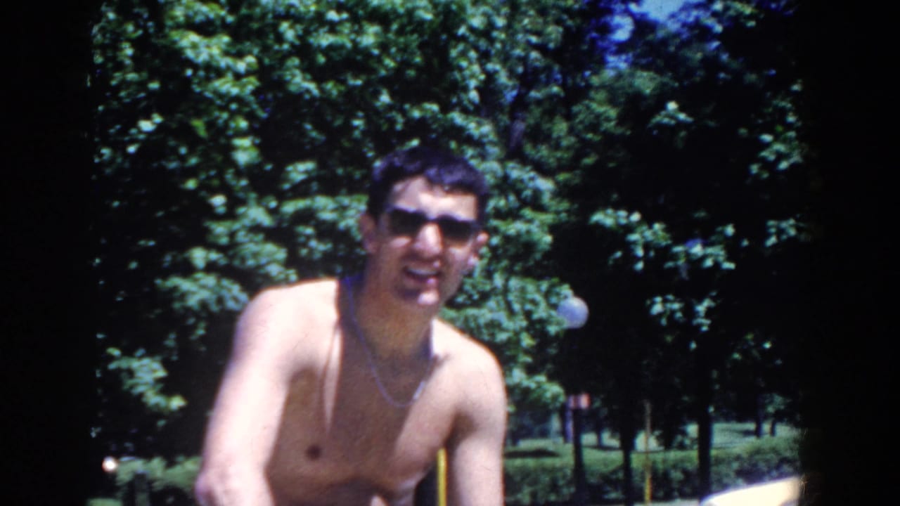 Shirtless man in sunglasses faces the camera with trees behind him.