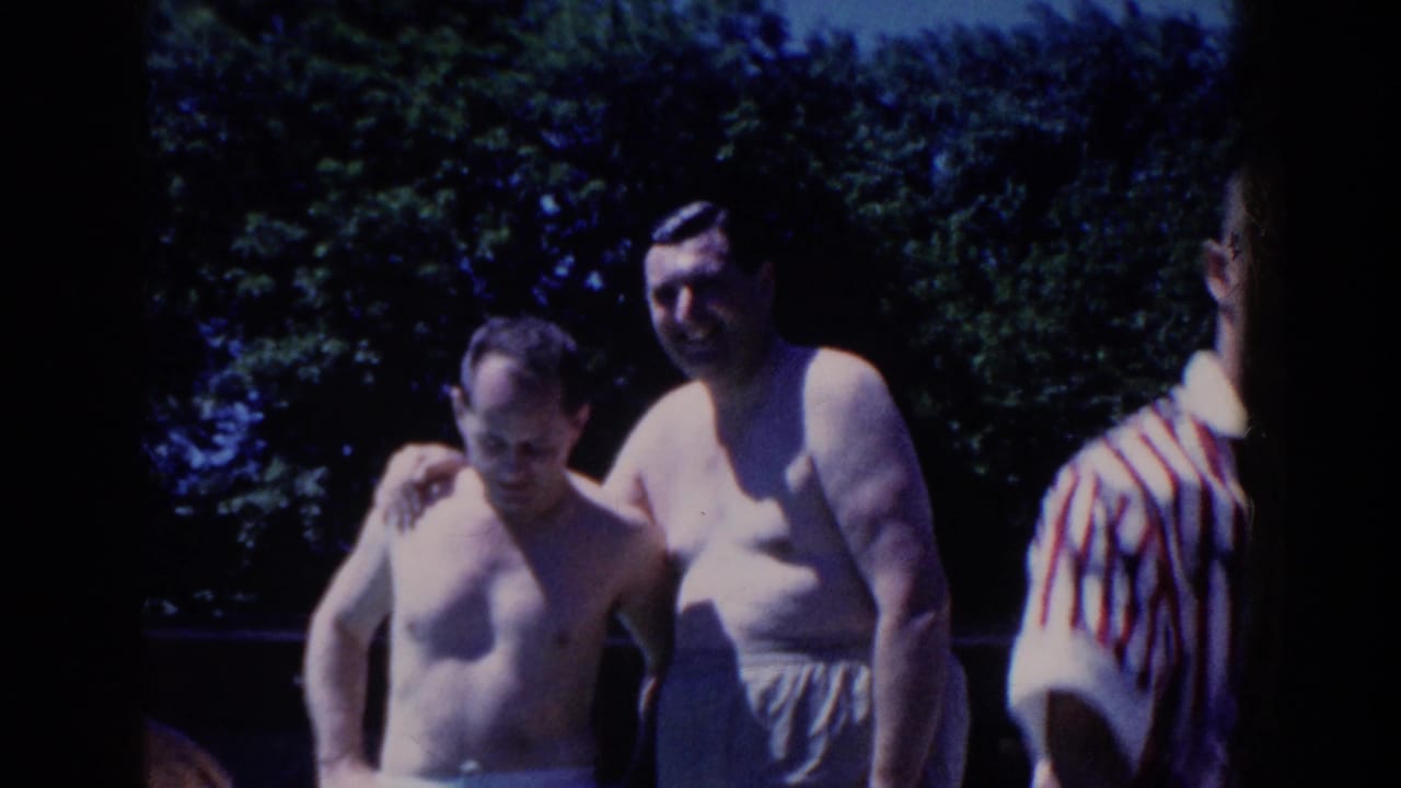 Two shirtless men stand together near trees at an outdoor swimming pool.