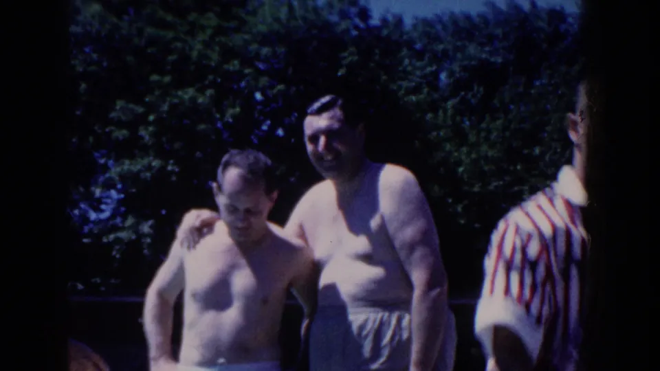 Two adult men in swimwear stand close together outdoors, with trees behind them at the French Lick swimming pool.