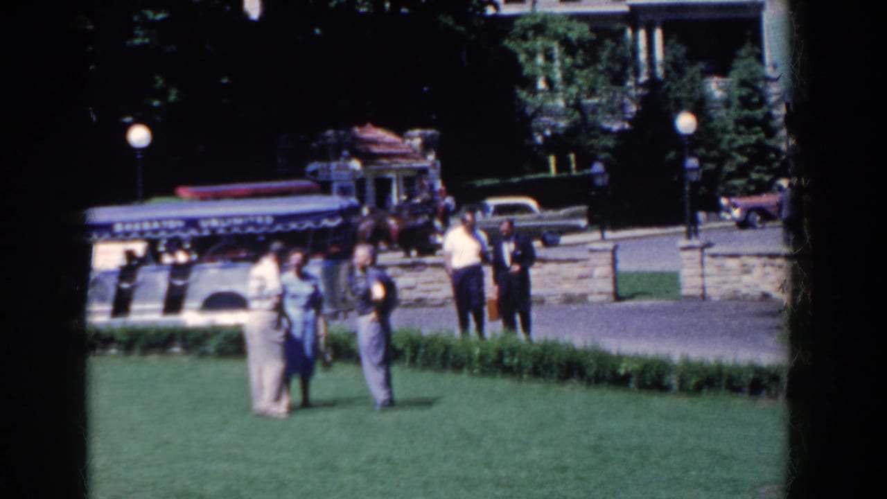 In French Lick, Indiana, adults in 1950s clothing talk on a manicured lawn as classic cars and a horse-drawn carriage pass behind them.
