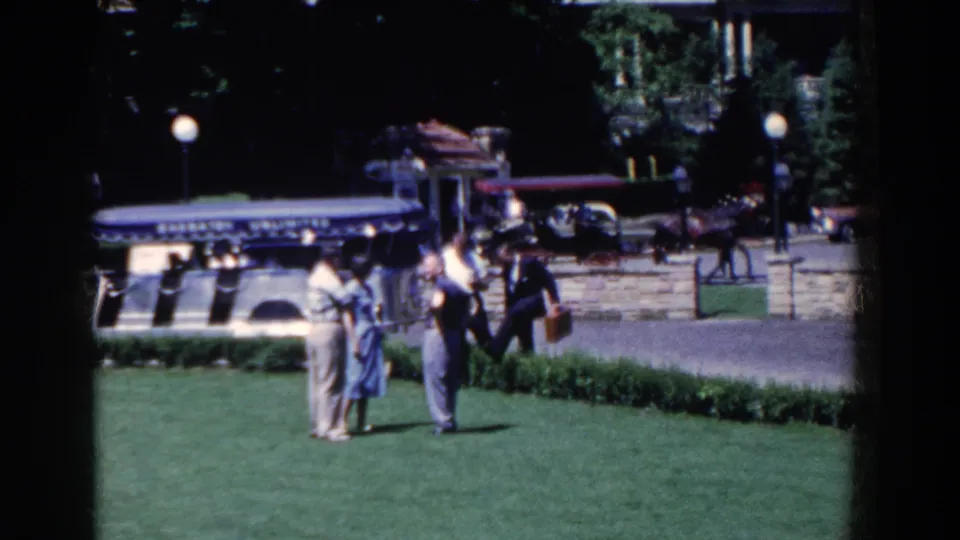 Adults in mid-century attire continue talking on a French Lick lawn, with a dark automobile, carriage, and stone wall in the background.