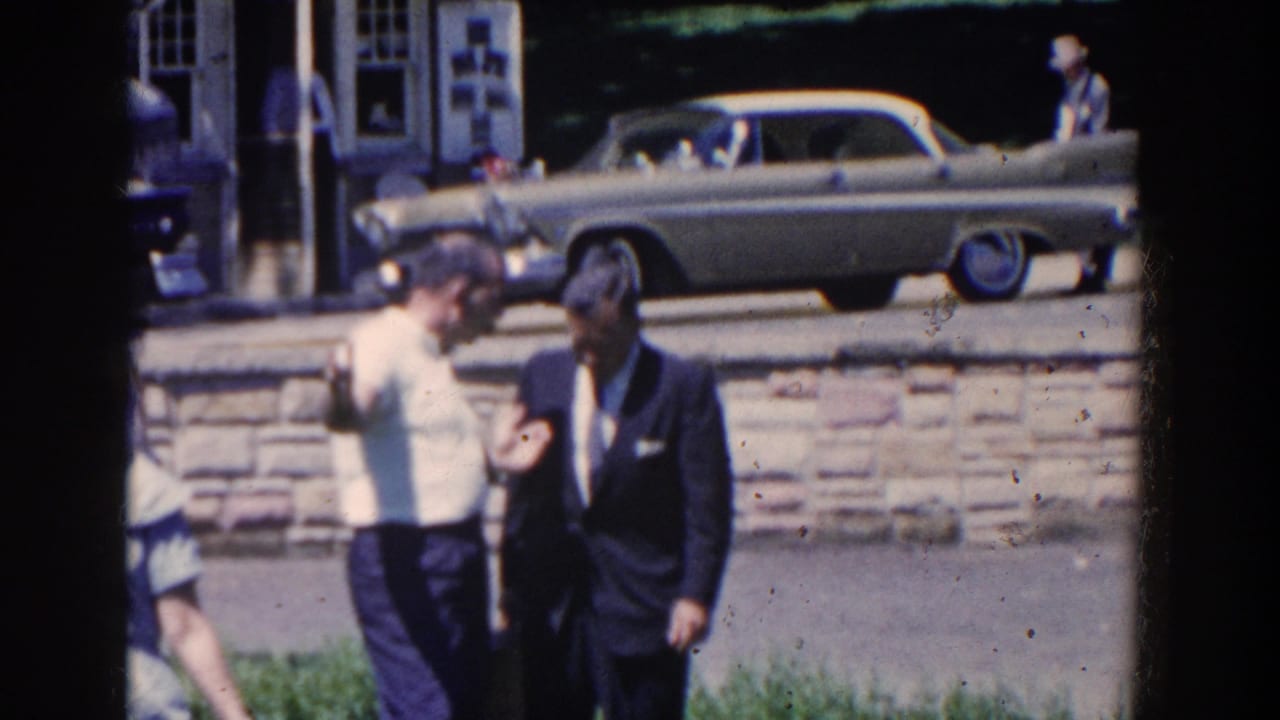 Two men stand near a white stone wall with a two-tone car parked behind them.