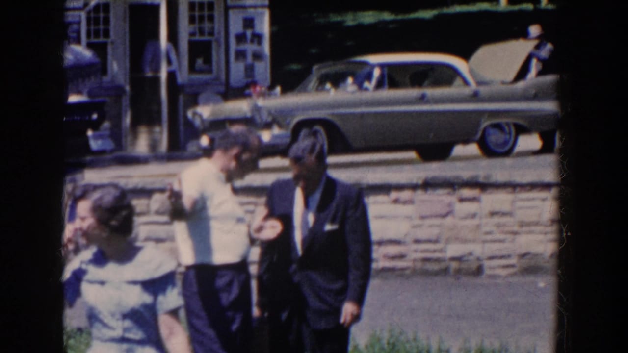 Pedestrians pass in front of two men, with a parked two-tone car behind a stone wall.
