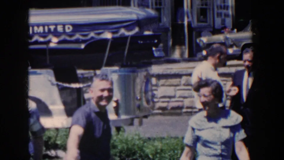 In the French Lick home-movie scene, a man and woman pass close to the camera while partial signage, a stone wall, and a parked car appear behind them.