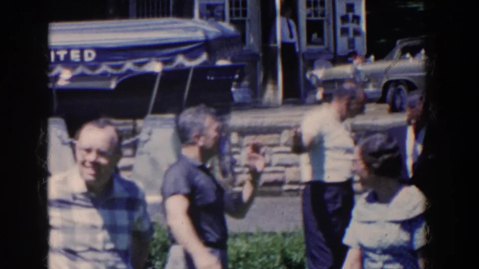 Several adults move through the sunny 1958 French Lick street scene, with a partial awning sign, white stone wall, and parked car visible behind them.