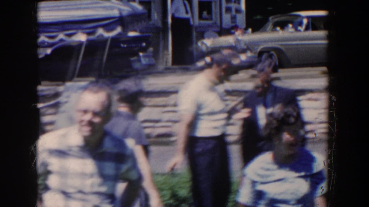 1958 French Lick home-movie frame with adults near a stone wall, sunlit walkway and parked mid-century cars.