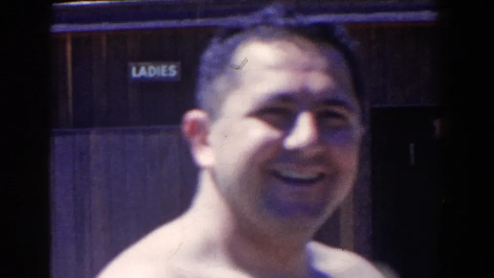 Shirtless man smiles toward the camera in 1958 French Lick footage, with a ladies sign and dark wooden doorway behind him.