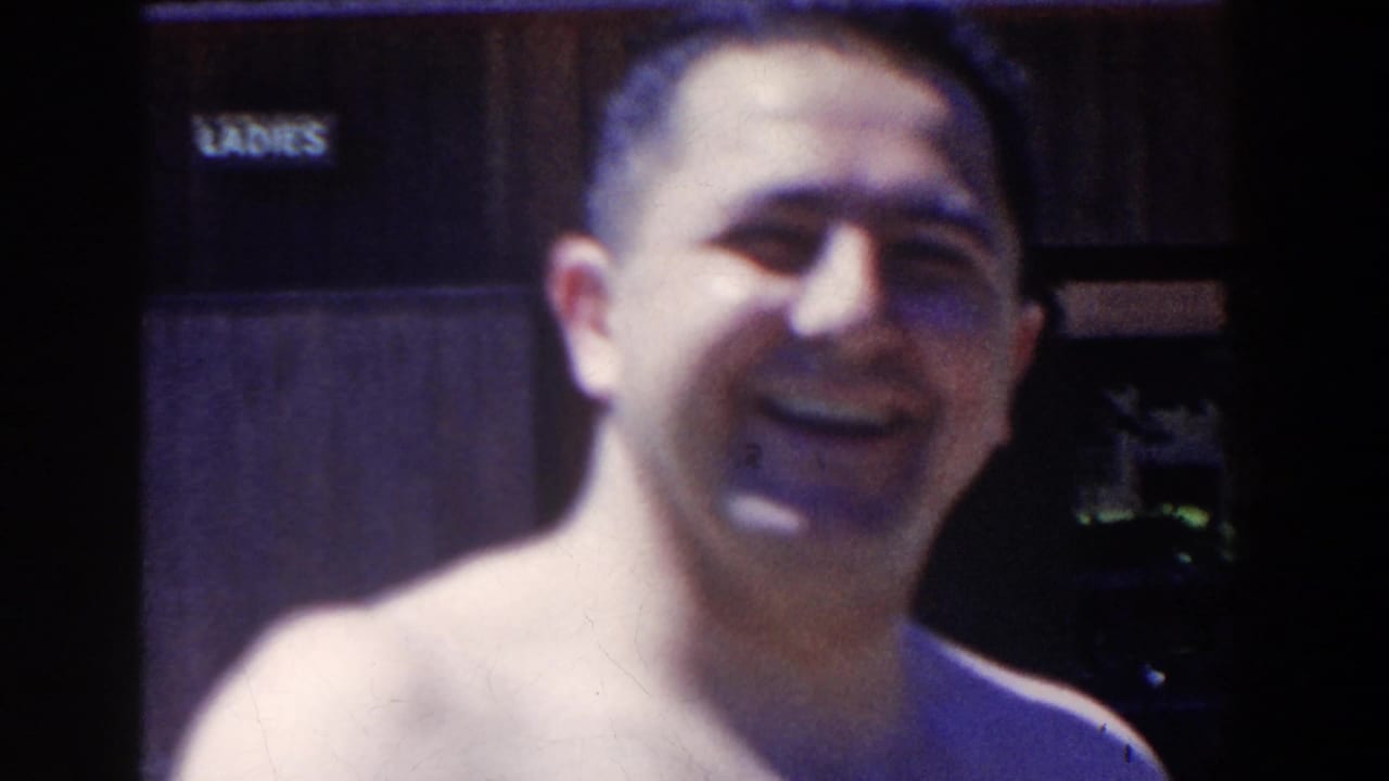 In 1958 French Lick, Indiana home-movie footage, a shirtless adult man smiles toward the camera near a LADIES restroom sign.