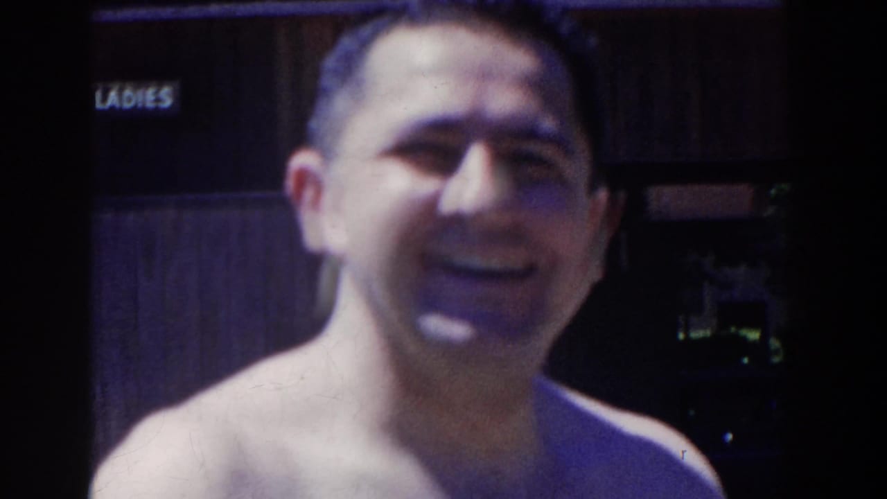 The adult man faces the camera in soft 8mm footage, with the LADIES sign and a shadowed doorway visible behind him.