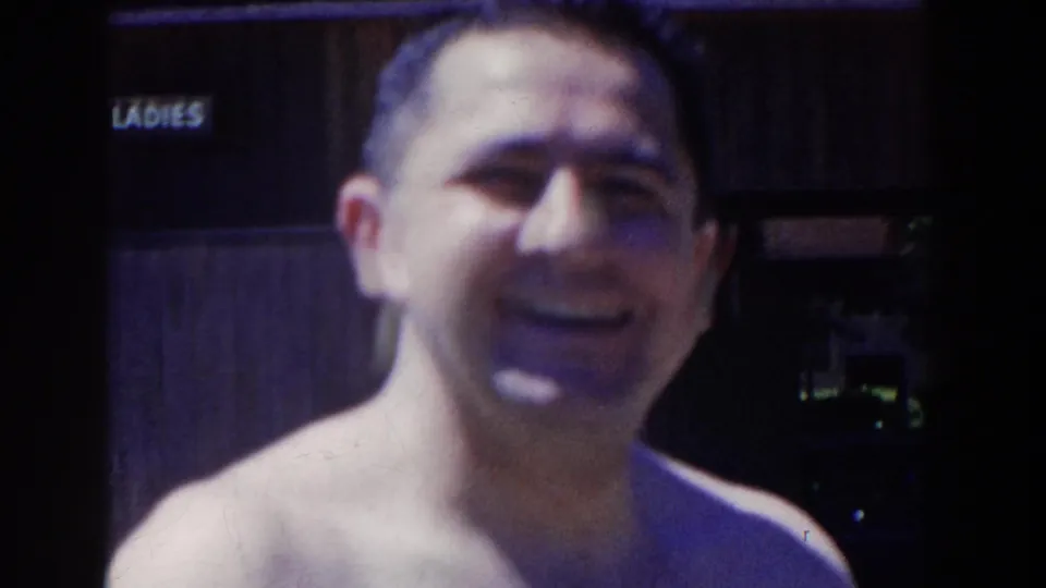 The adult man faces the camera in soft 8mm footage, with the LADIES sign and a shadowed doorway visible behind him.