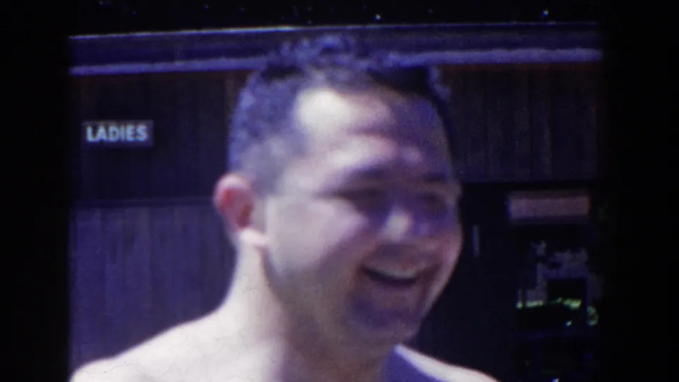 The man turns partly sideways and laughs, captured in grainy color 8mm beside the visible LADIES sign.