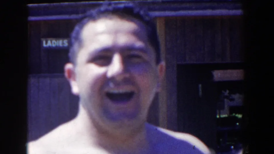 The adult man laughs openly toward the camera in the French Lick home movie, with the LADIES sign behind him.
