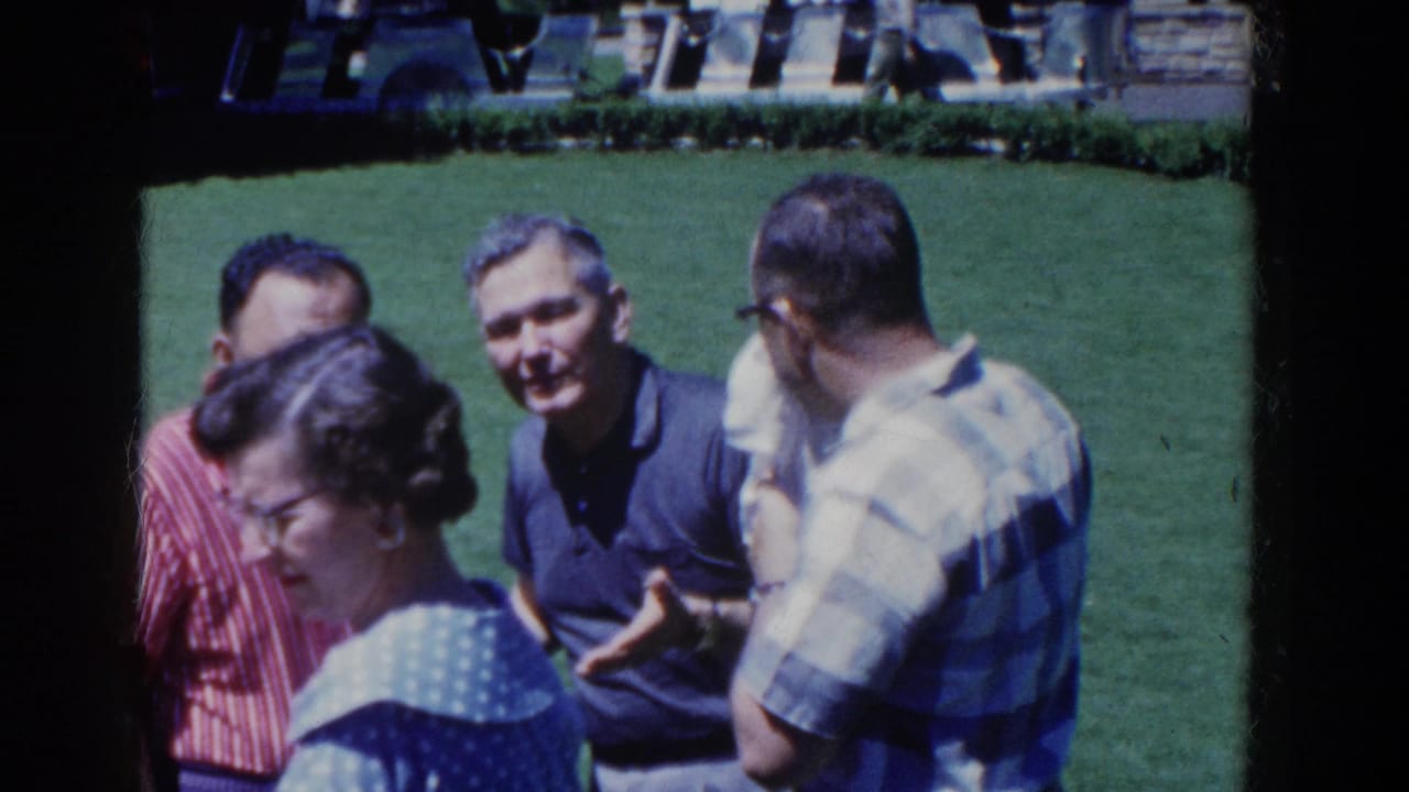 1958 French Lick frame showing adults clustered on a sunlit lawn, with faces visible and casual short-sleeve clothing.