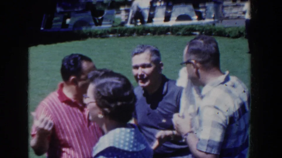 1958 French Lick home-movie still of adults talking on a lawn, with a smiling man, a red striped shirt, and a woman in the foreground.