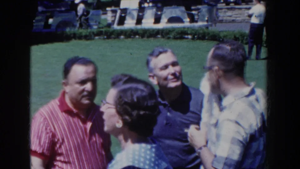 1958 French Lick frame of four adults gathered outside on grass, with landscaped plantings behind them and visible mid-century casual clothing.