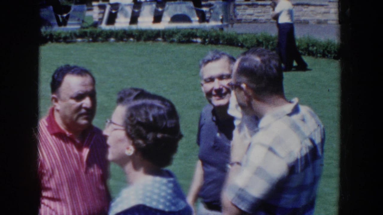 1958 French Lick home-movie frame showing adults in casual clothing conversing on a green lawn near stone landscaping.