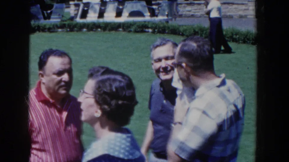 1958 French Lick home-movie frame showing adults in casual clothing conversing on a green lawn near stone landscaping.