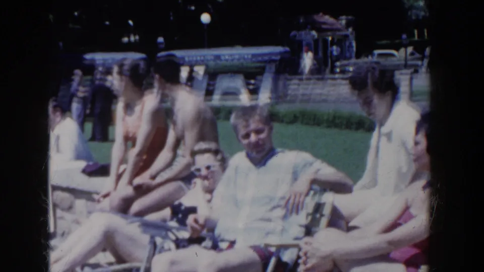 French Lick home-movie frame of adults seated on folding lawn chairs, with grass, stonework, and parked cars behind them.