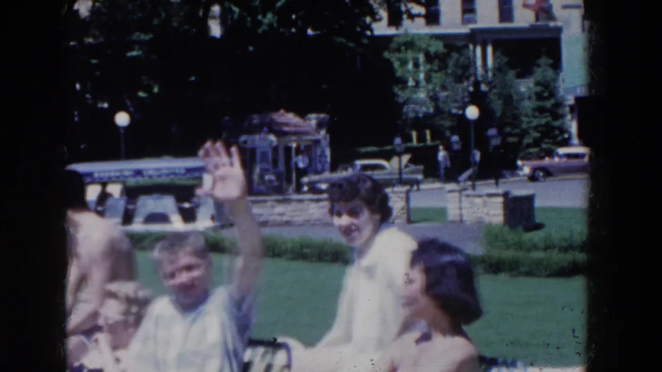 A man in a blue shirt waves toward the 8mm camera while seated with friends outdoors in 1958 French Lick, Indiana.