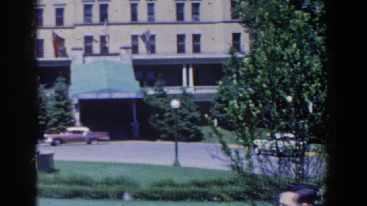 1958 French Lick hotel exterior with a green awning, landscaped lawn, trees, lamps, and a pink car near the entrance.