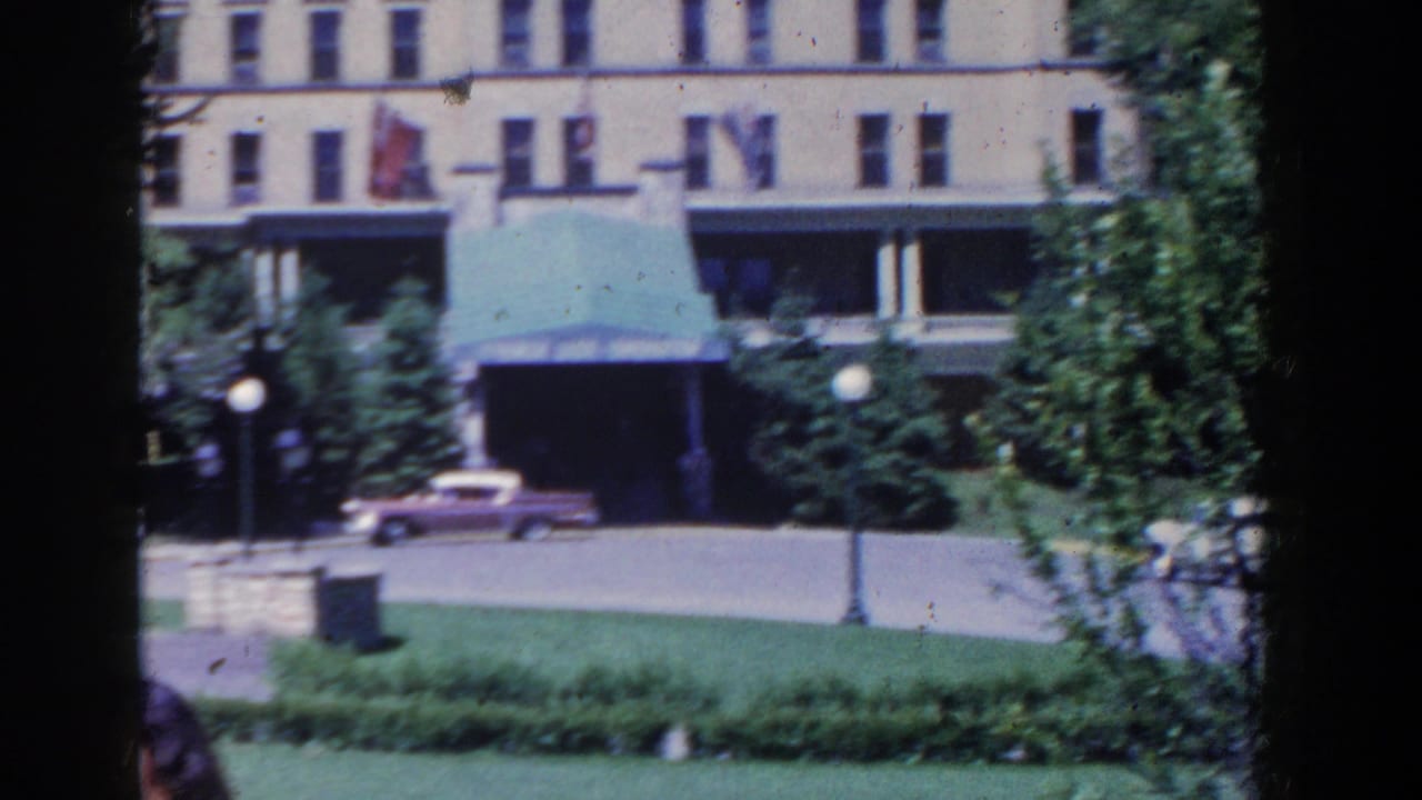 French Lick hotel exterior in 1958, framed by trees and lawn, with a green awning, street lamps, and a pink car.