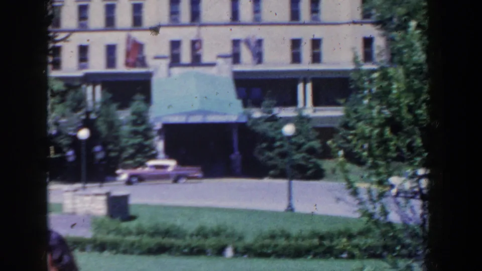 French Lick hotel exterior in 1958, framed by trees and lawn, with a green awning, street lamps, and a pink car.