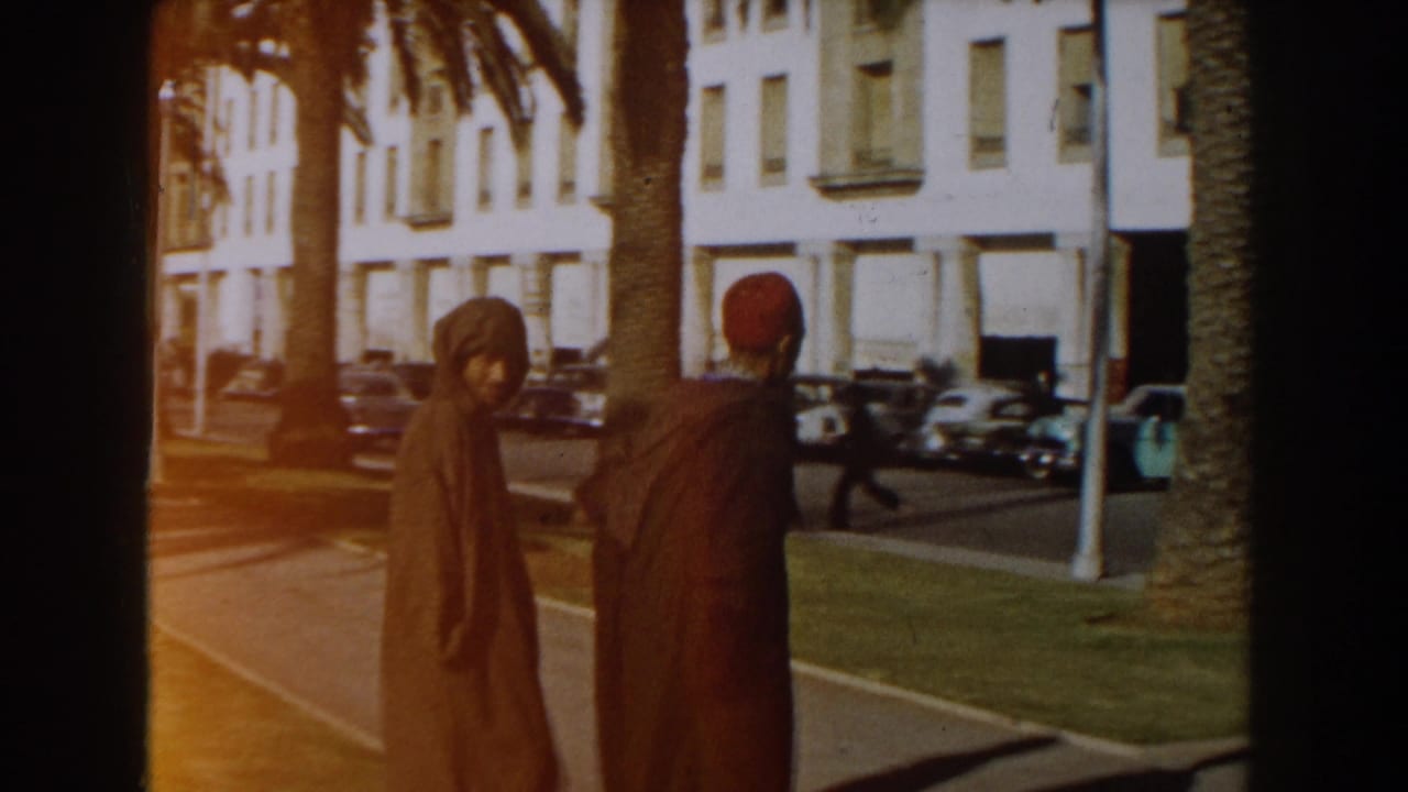 Two men in robes walk on a sunny sidewalk beside palm trees and parked cars.