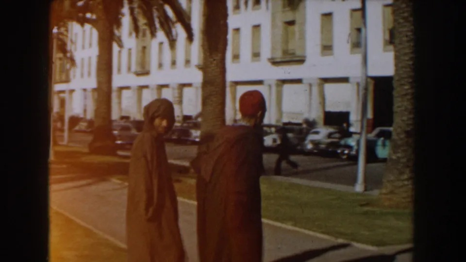 1959 Casablanca home-movie frame showing two men in traditional attire walking near palm trees, parked cars, and white buildings.