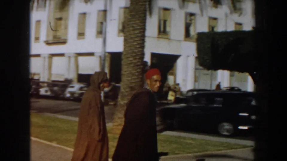 1959 Casablanca street frame with a man in a red fez and another in a hooded robe beside traffic and palm-lined buildings.