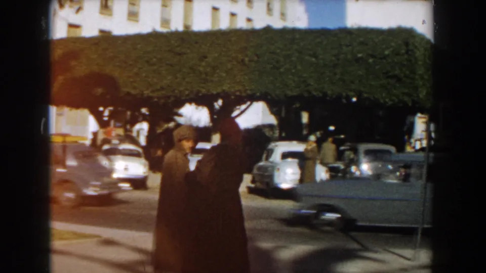 1959 Casablanca frame showing two men near a curb, passing traffic, parked cars, and clipped street trees in bright daylight.
