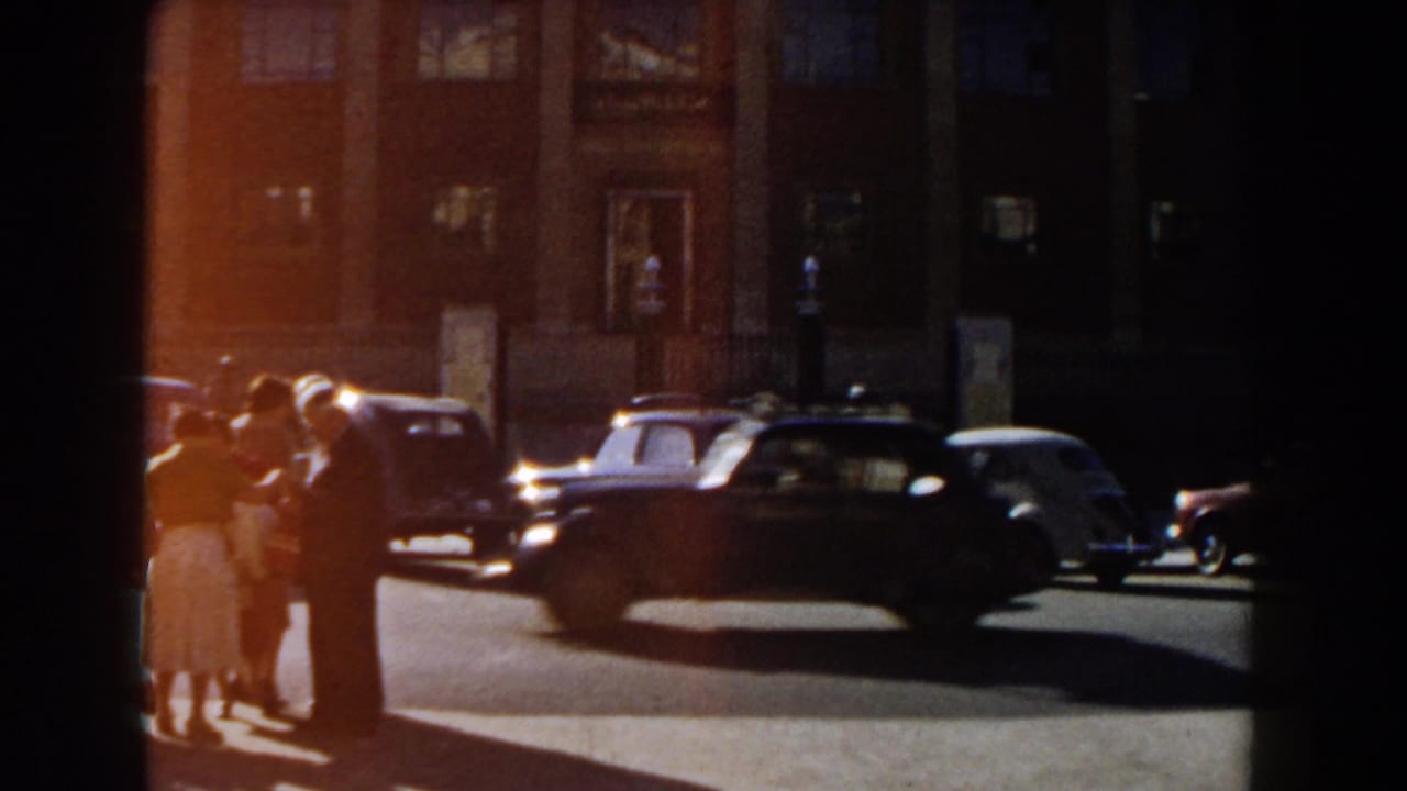Pedestrians stand near parked and passing cars outside a large building with windows.