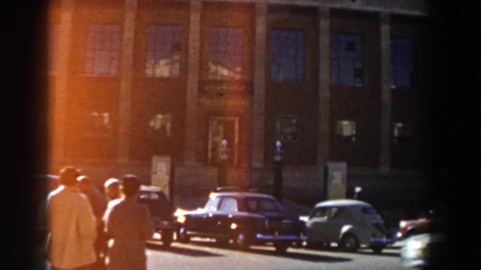 1959 Casablanca view of pedestrians beside parked and moving cars, with tall windows and pillars on the building behind them.