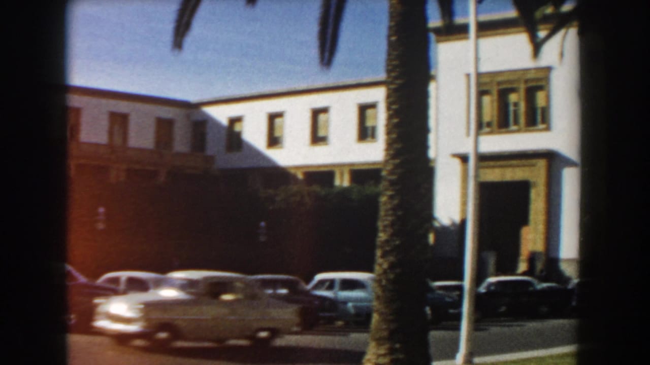 1959 Casablanca street view with a long white building, palm trunks, flagpoles, parked cars, and a passing light-colored car.