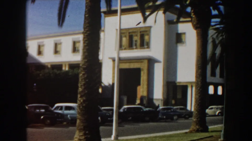White Casablanca building seen beyond palm trunks and flagpoles, with several mid-century cars parked or moving along the street.