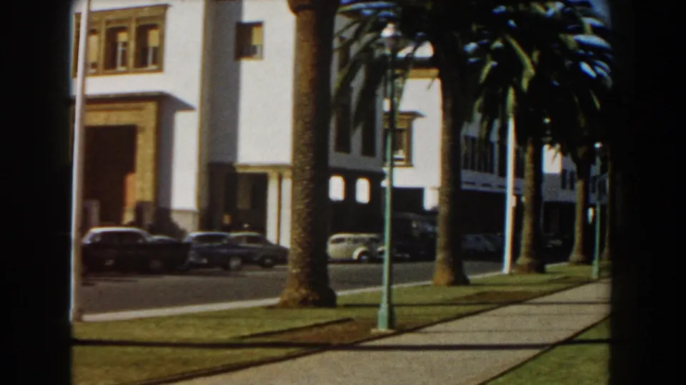 Casablanca exterior with a sidewalk, palms, flagpoles, a white building entrance, and parked cars along the sunny street.