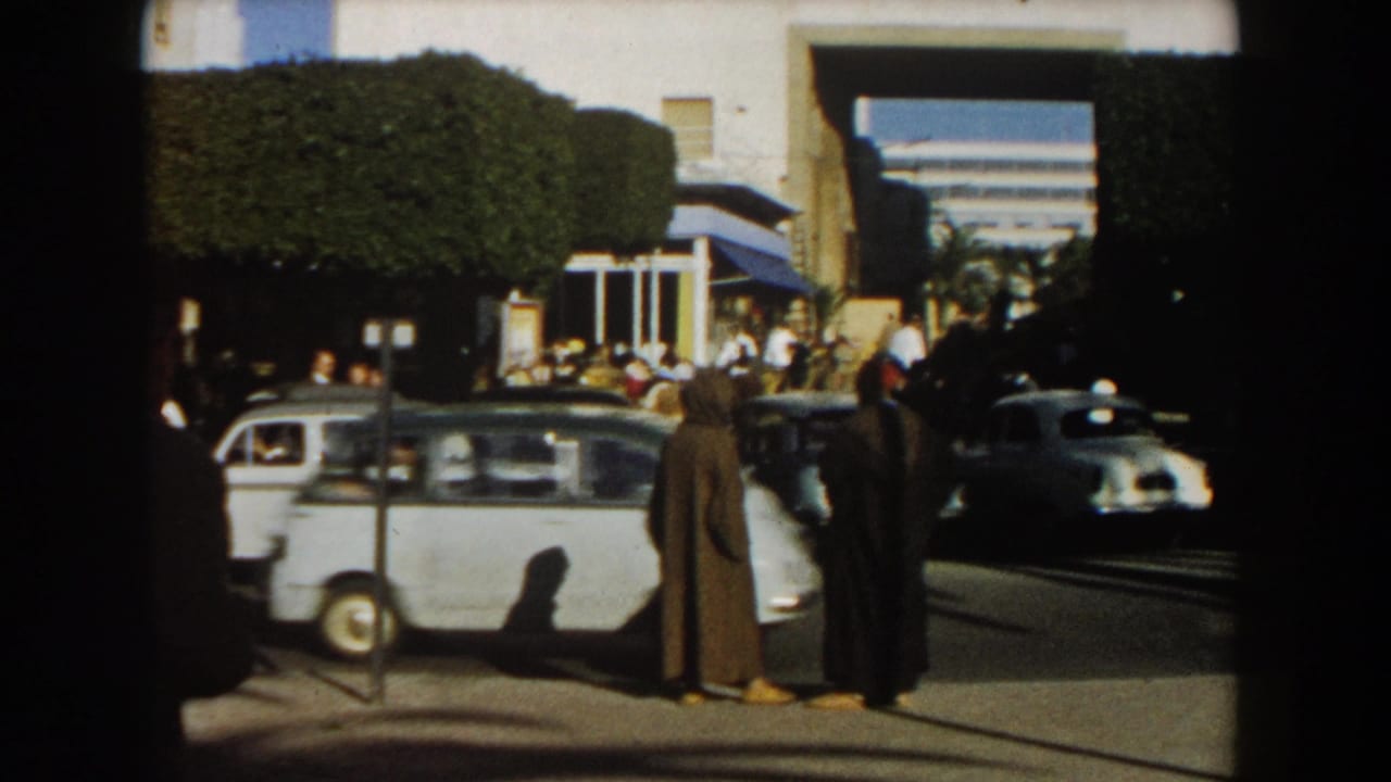 1959 Casablanca sidewalk scene with two men in long cloaks standing near traffic, parked cars, and a crowd by white buildings.