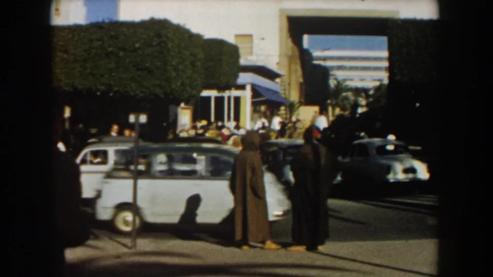 1959 Casablanca sidewalk scene with two men in long cloaks standing near traffic, parked cars, and a crowd by white buildings.