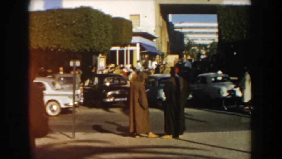1959 Casablanca street corner with cloaked men in the foreground, pedestrians across the sidewalk, and several mid-century cars nearby.