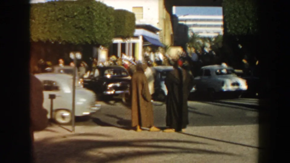 1959 Casablanca street view with two cloaked men seen from behind, cars moving and parked nearby, and pedestrians outside white buildings.