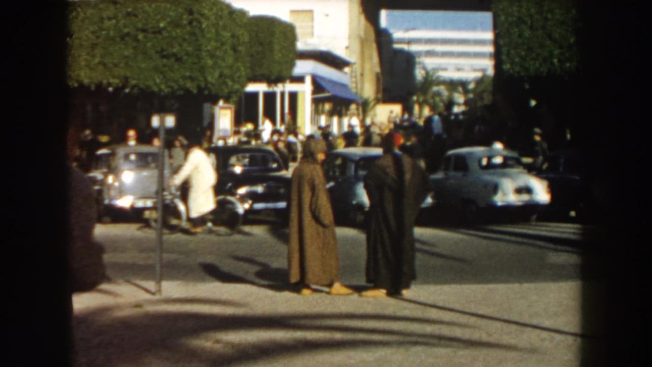 In 1959 Casablanca, two robed men stand near the curb as mid-century cars pass and pedestrians gather by hedges and a building entrance.