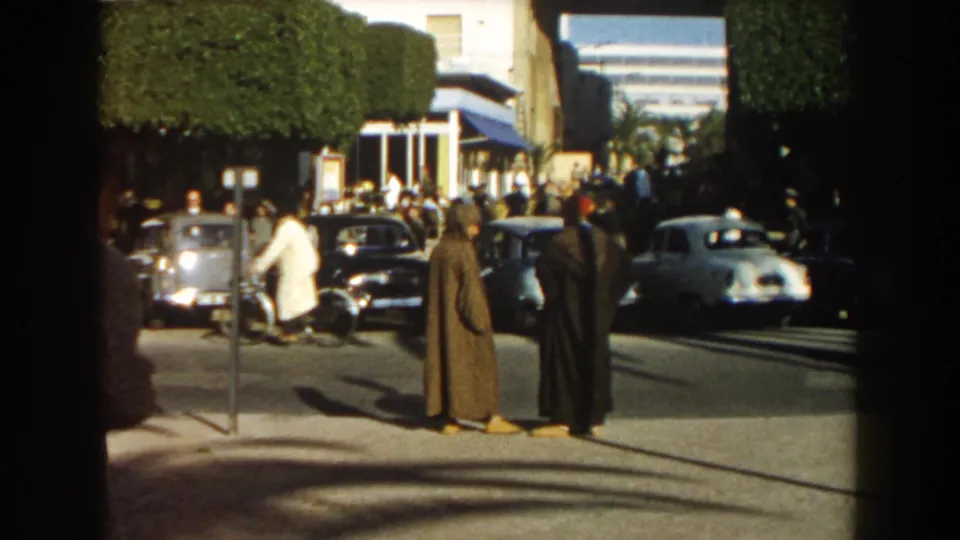 In 1959 Casablanca, two robed men stand near the curb as mid-century cars pass and pedestrians gather by hedges and a building entrance.