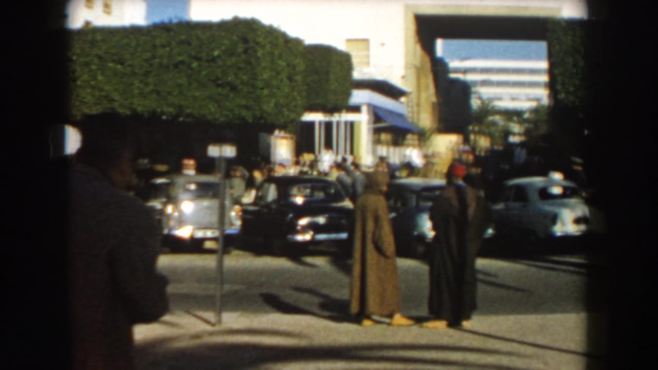 In Casablanca in 1959, two men in long robes stand in conversation while dark and light 1950s cars move through the intersection.