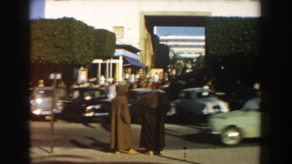 A light-colored car passes in motion blur in front of two robed men on a 1959 Casablanca street with pedestrians and hedges behind them.