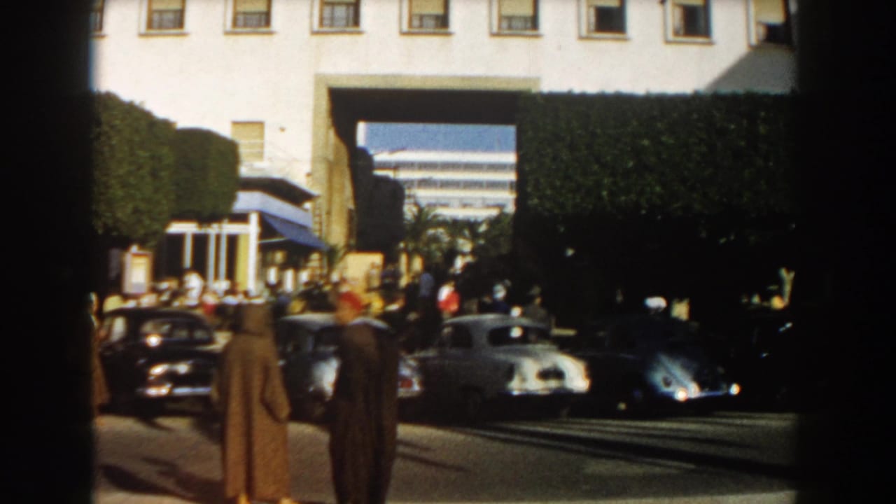 1959 Casablanca street view with classic cars, pedestrians in period dress, clipped hedges, and a white building with a prominent archway.