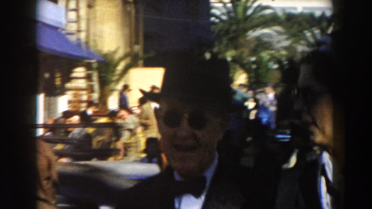 Elderly man in a dark suit and fedora walks on a busy sunlit street.