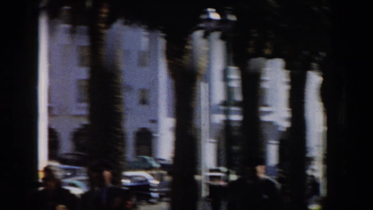 1959 Casablanca home-movie frame showing palm trunks, white building facades, parked period cars, and shaded pedestrians.