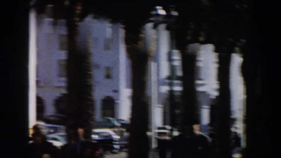 1959 Casablanca home-movie frame showing palm trunks, white building facades, parked period cars, and shaded pedestrians.